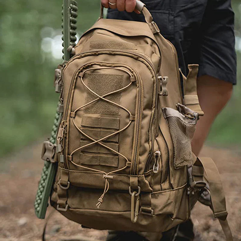 Tan backpack with multiple pockets and straps held by a person in a forest setting